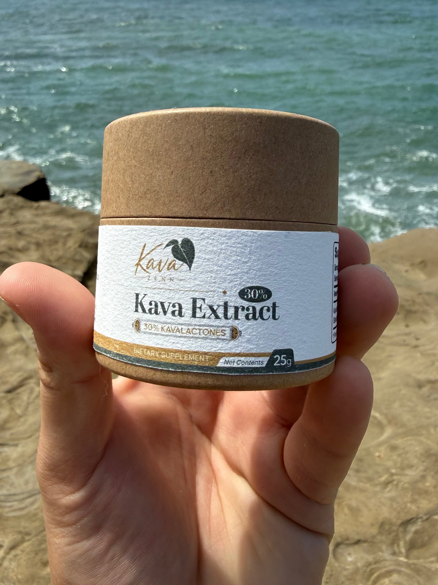 A hand holding a small jar of Kava Extract against a beach background with ocean water and rocks.
