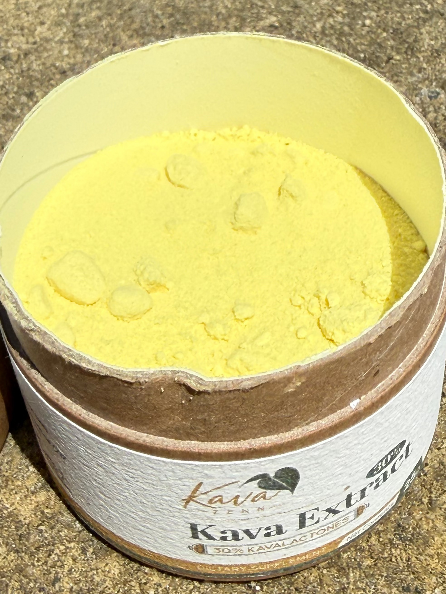 An open round cardboard tin of Kava Extra coffee powder, showing the pale yellow powder inside, placed on a textured concrete surface outdoors.